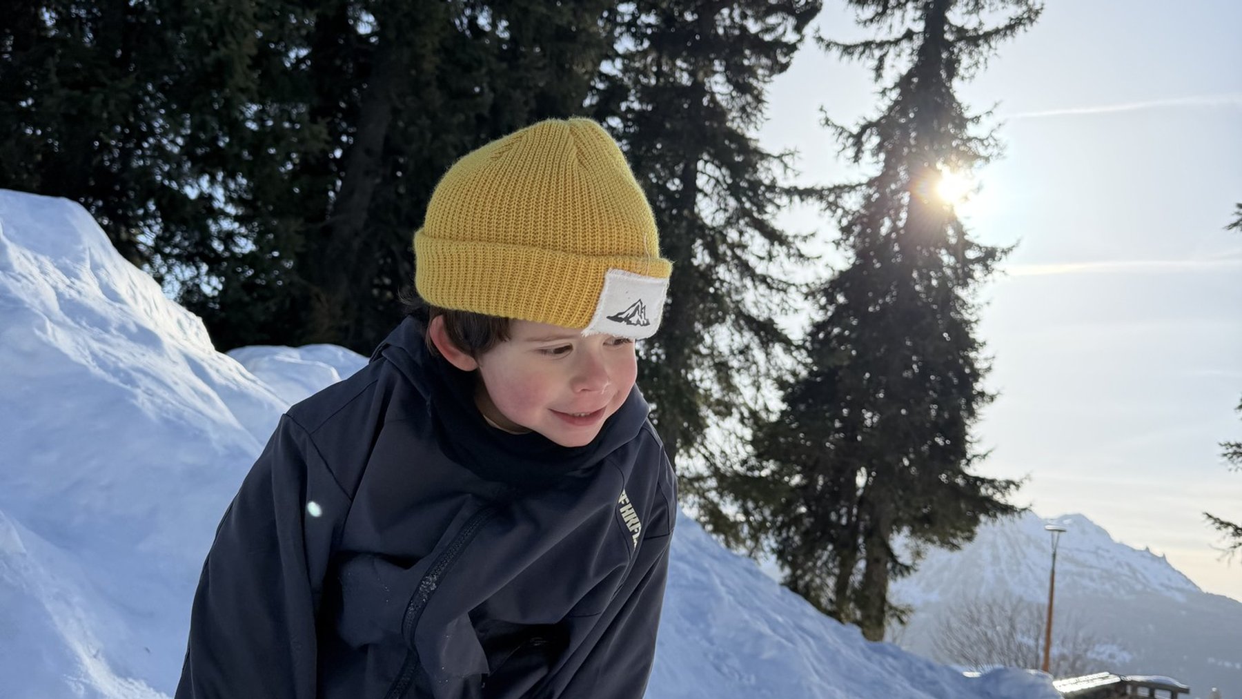 Snow-suited child in a mustard beanie and jacket standing on a sunny ski slope in alpine trees