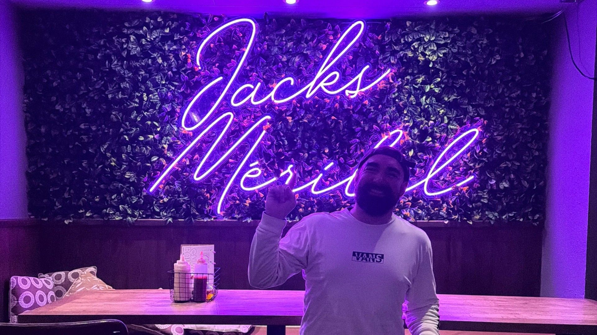A man stands inside Jack’s beneath a glowing neon sign, with a bar and purple lighting behind him.