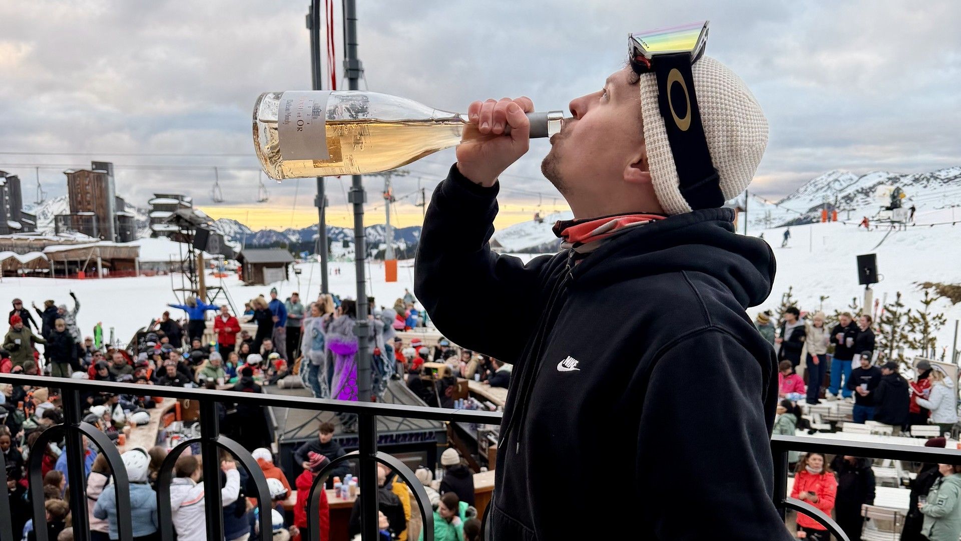 Ski fan cheers at a crowded outdoor event, drinking from a bottle on the snow