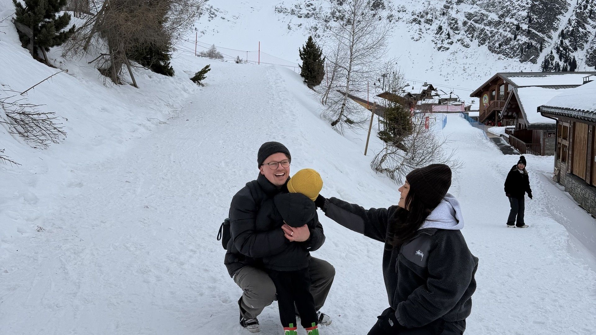 Two people kneel on a snowy path in La Rosière, with chalets and snow-covered mountains behind them.