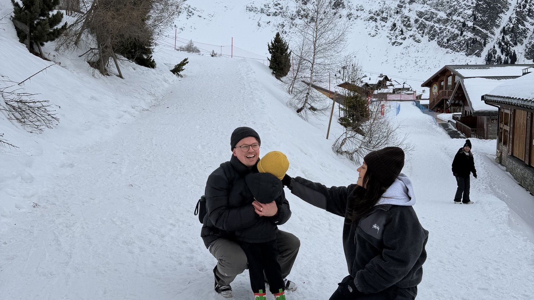 Two people kneel on a snowy path in La Rosière, with chalets and snow-covered mountains behind them.