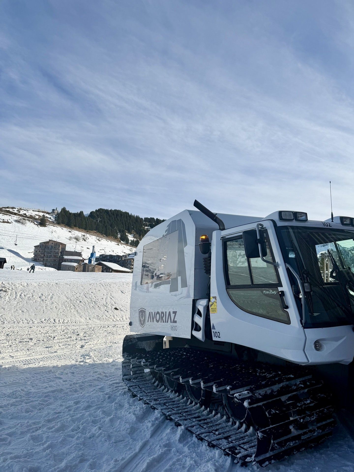 White tracked snow vehicle clearing snowy slopes with mountains and ski buildings in the background