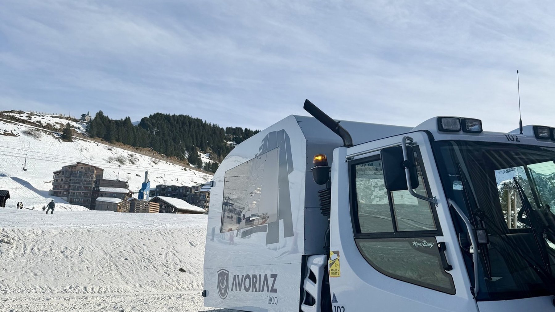 White tracked snow vehicle clearing snowy slopes with mountains and ski buildings in the background