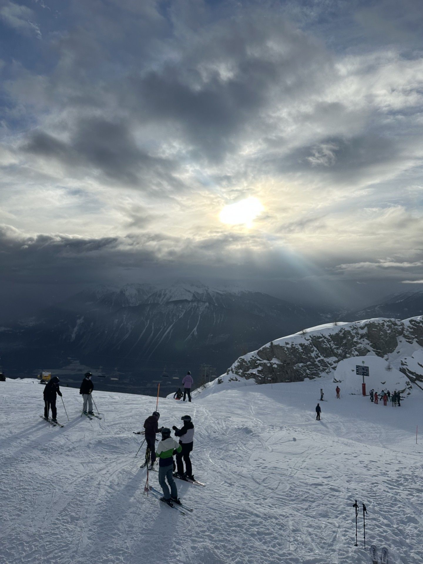 Skiers walk on a snowy mountain slope toward bright sun rays through clouds