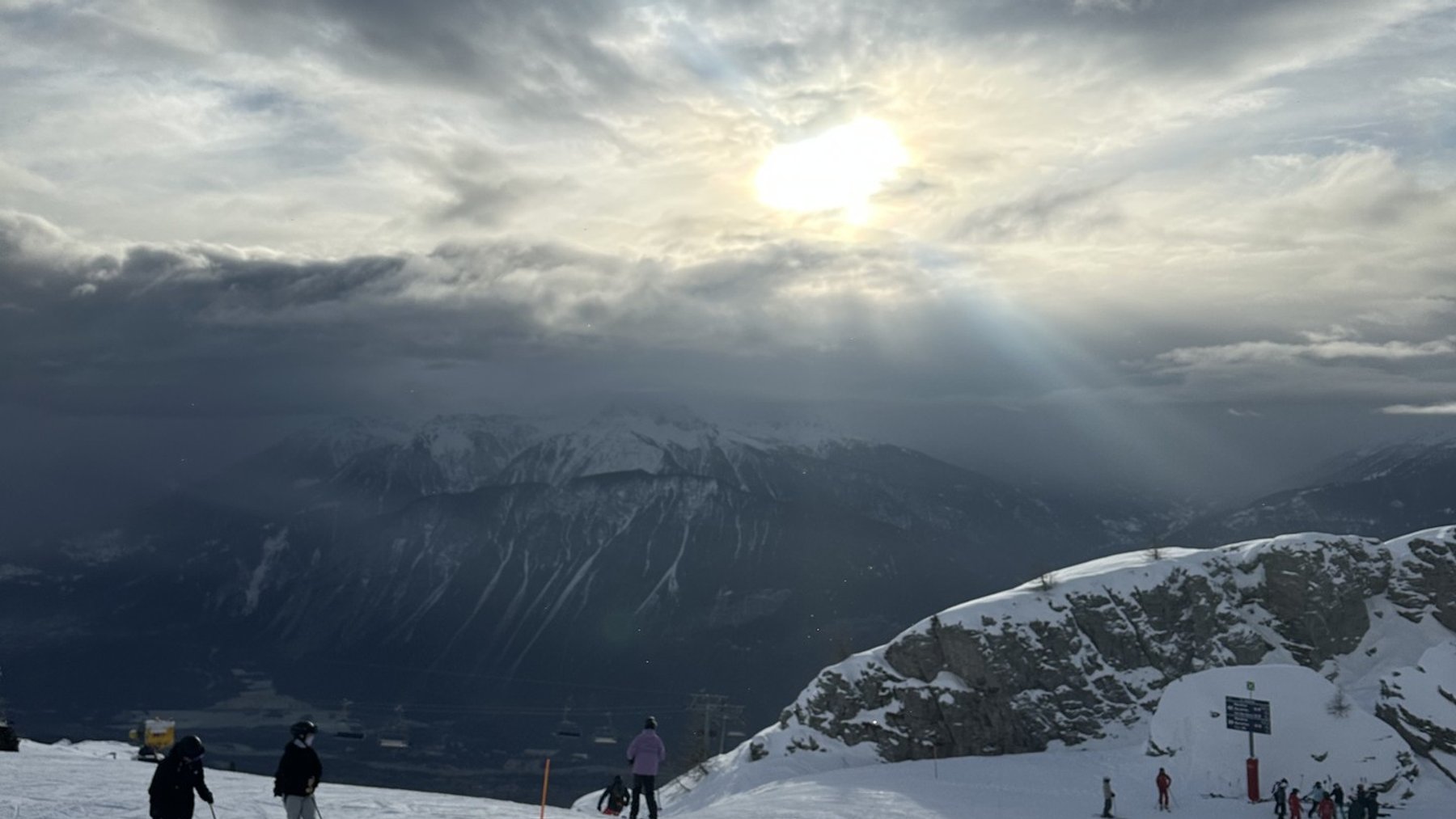 Skiers walk on a snowy mountain slope toward bright sun rays through clouds