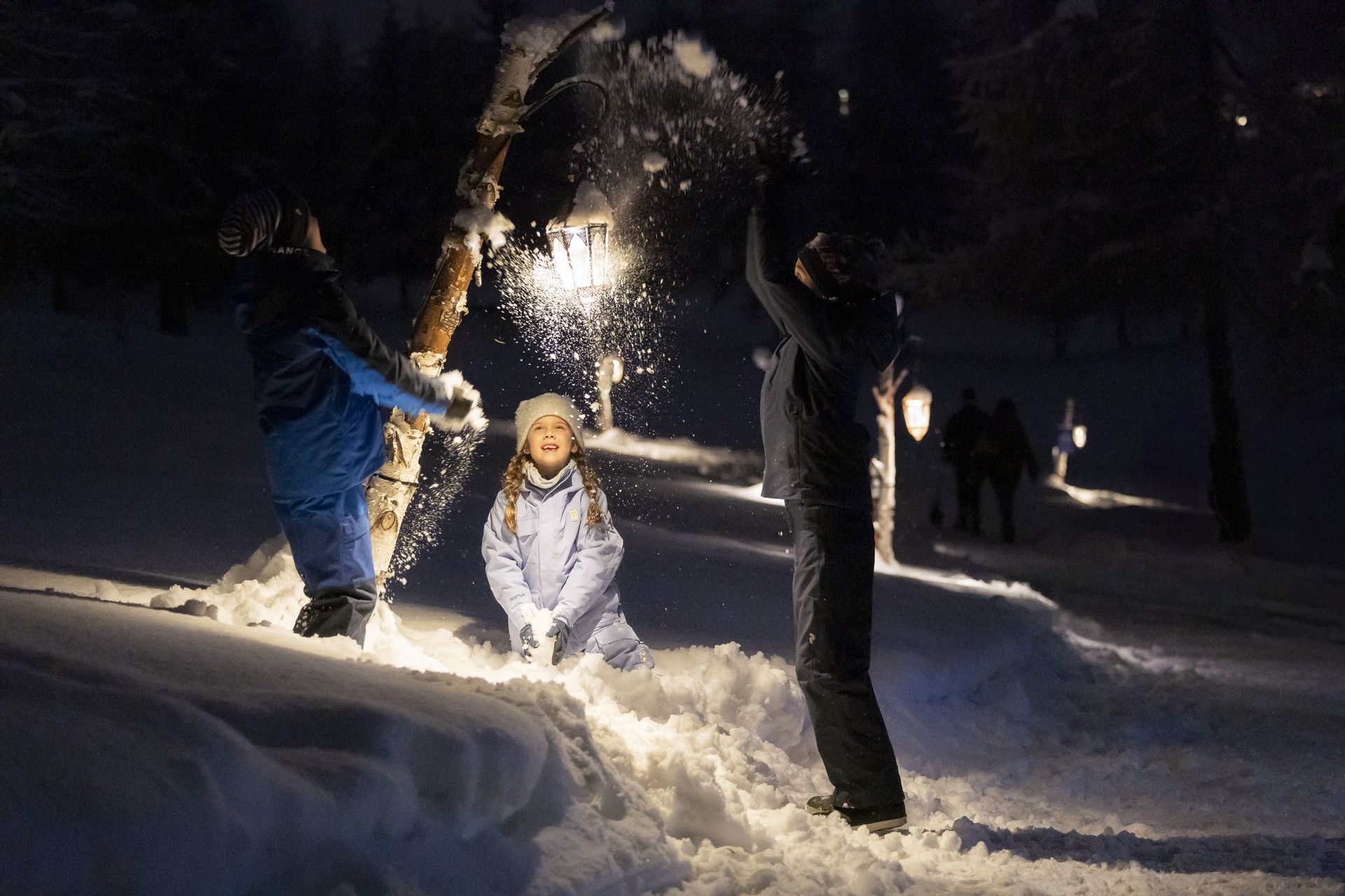 Family plays in fresh snow at night, throwing snow as holiday lights glow in the background