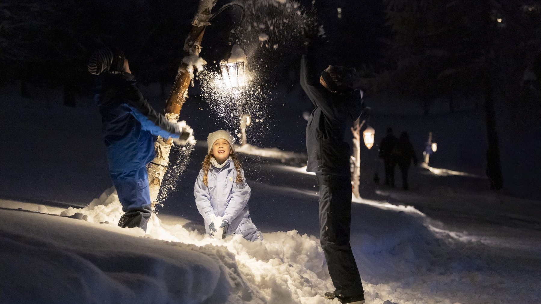 Family plays in fresh snow at night, throwing snow as holiday lights glow in the background