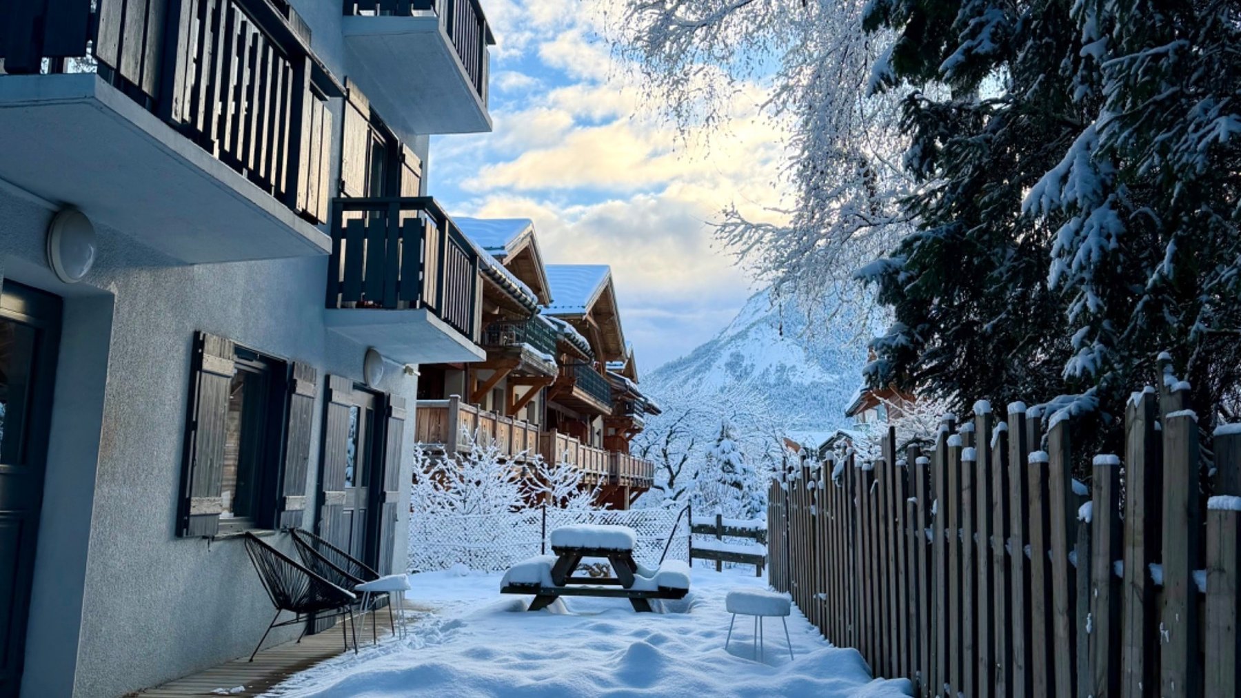 Snow-covered walkway beside chalet balconies and a wooden fence in Le Praz, with frosted trees and mountains beyond.