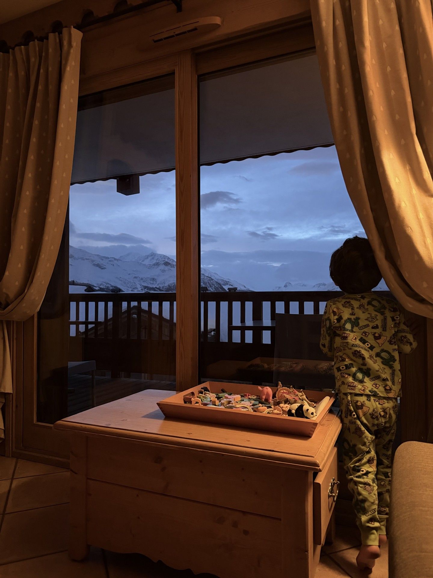 View through an open window at a ski lodge, with curtains framing snowy mountain peaks.
