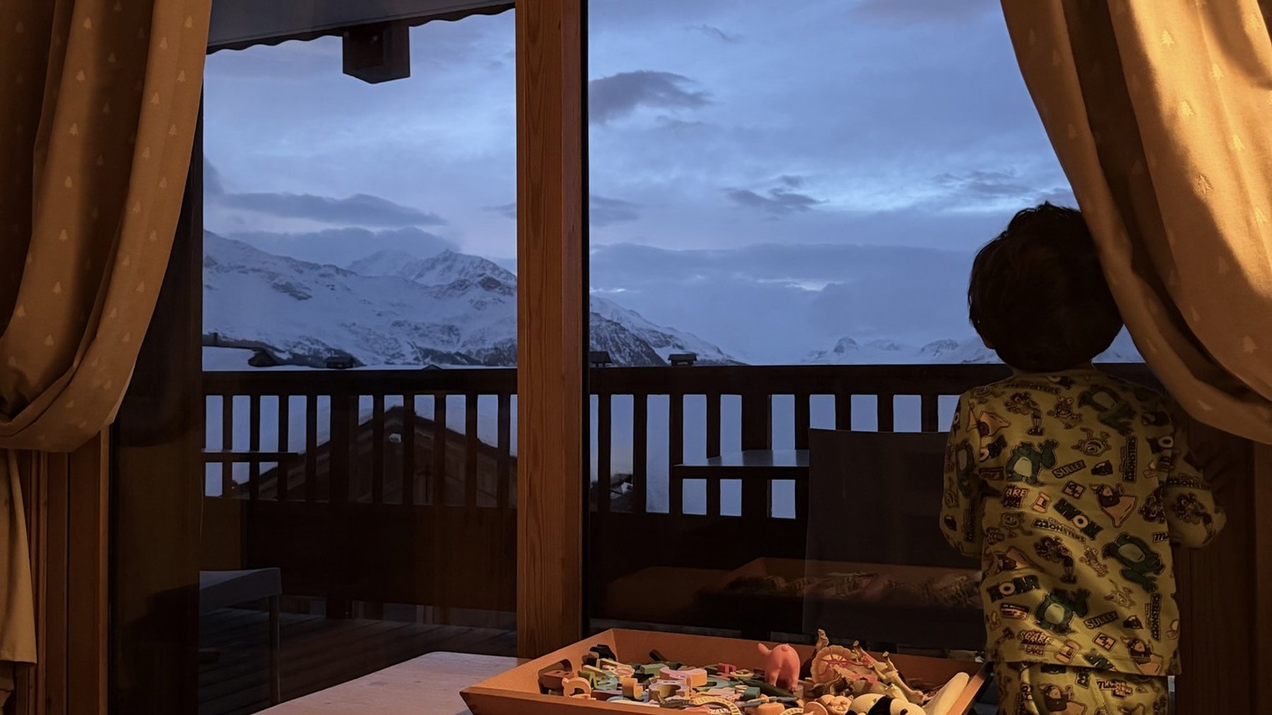 View through an open window at a ski lodge, with curtains framing snowy mountain peaks.