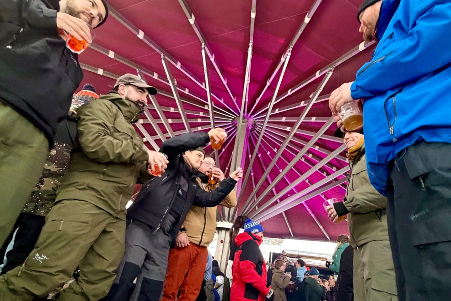 Skiers in winter clothing gather under a red canopy for drinks at an après-ski event