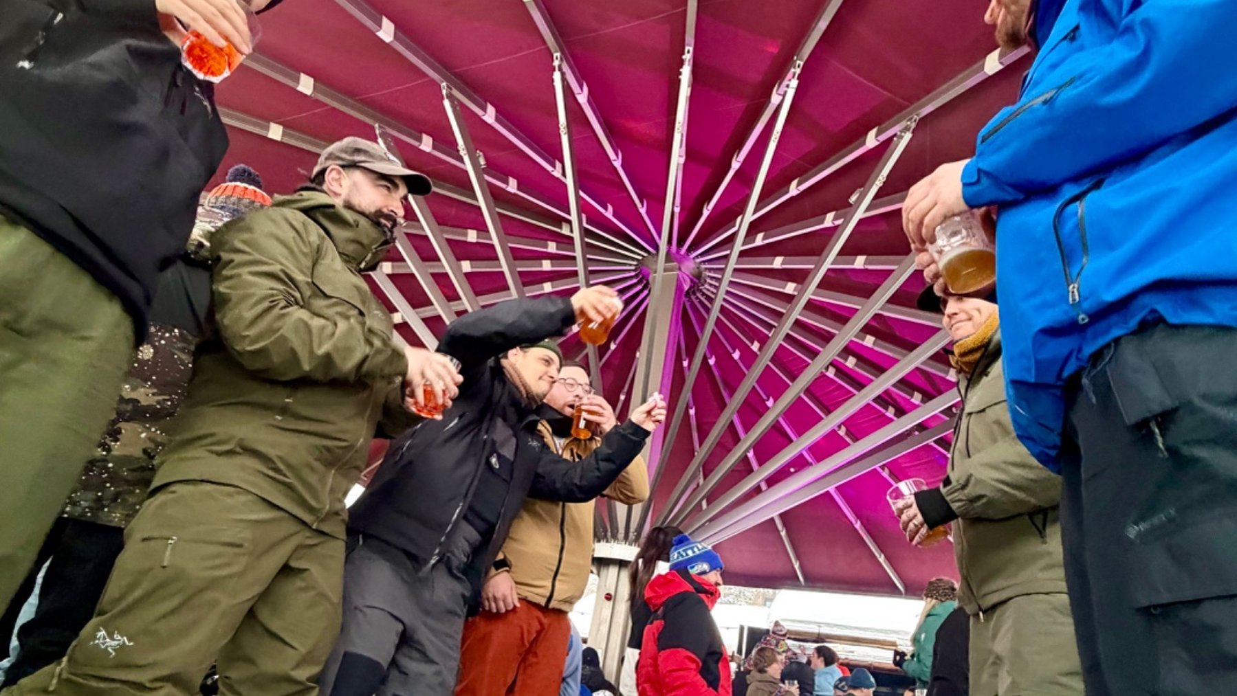 Skiers in winter clothing gather under a red canopy for drinks at an après-ski event