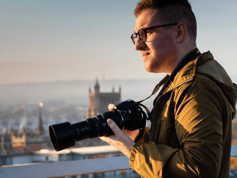 RSPB Big Garden Bird Watch campaign - Man wearing glasses stood on the roof of a building overlooking a city holding a professional camera with long lens as the sun is rising.