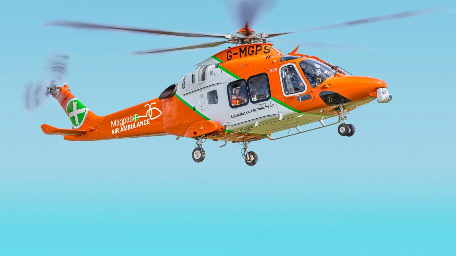 Bright orange Magpas helicopter in full view hovering in the air against a blue sky