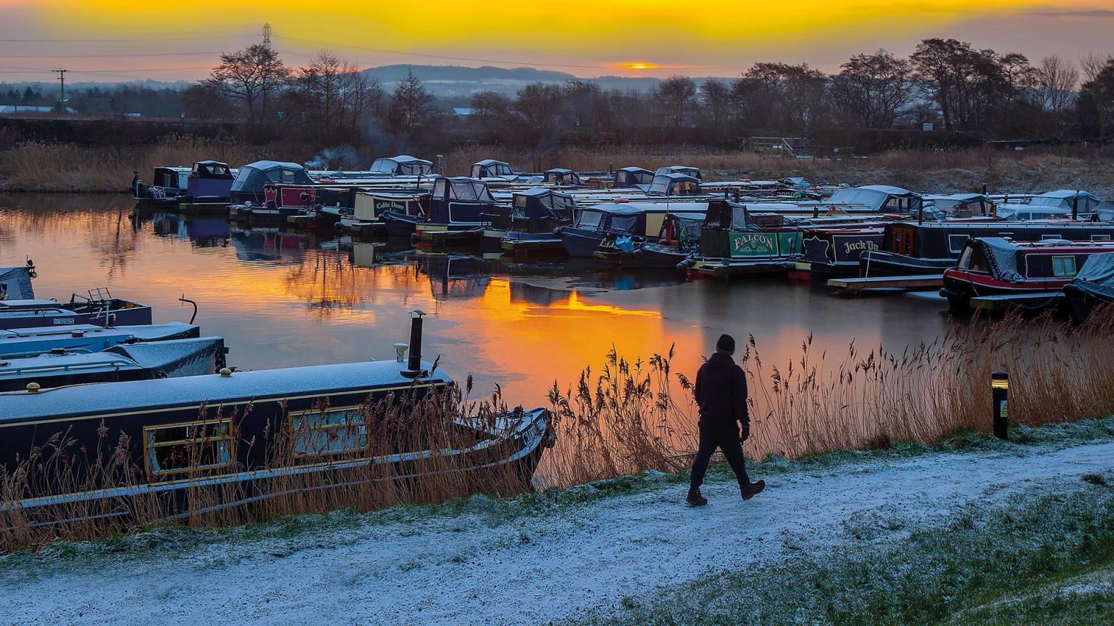 Winter sunset over a barge storage pond surrounded by brush land with a man walking to the right on a snow covered canal path. 