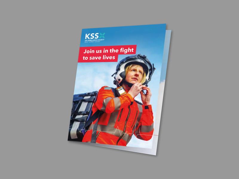 KSS Air Ambulance - I am 1 in 10 campaign mailer front cover displaying an image of a helicopter pilot ready for action wearing a medical supply chest on her back.