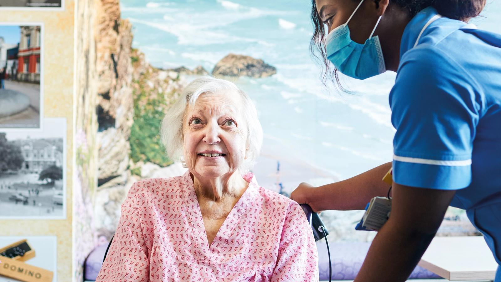 A seated elderly woman in a pink gown being given care by a nurse wearing a blue mask.