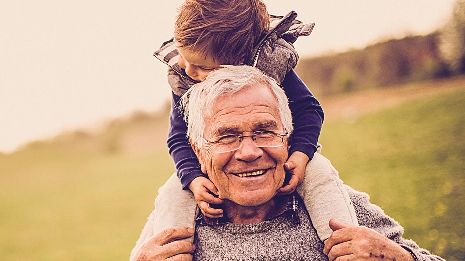 A relative close up of a smiling, old man wearing spectacles, walking in open countryside with a young child on his shoulders holding on.