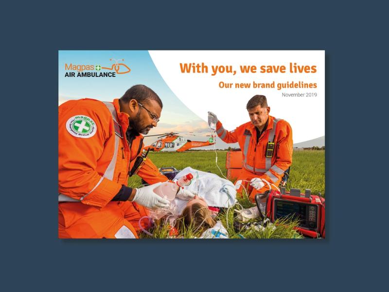 Magpas brand guidelines cover showing two male paramedics on the ground helping a patient with a bright orange helicopter in the background with the caption "With you, we save lives. Our new brand guidelines".