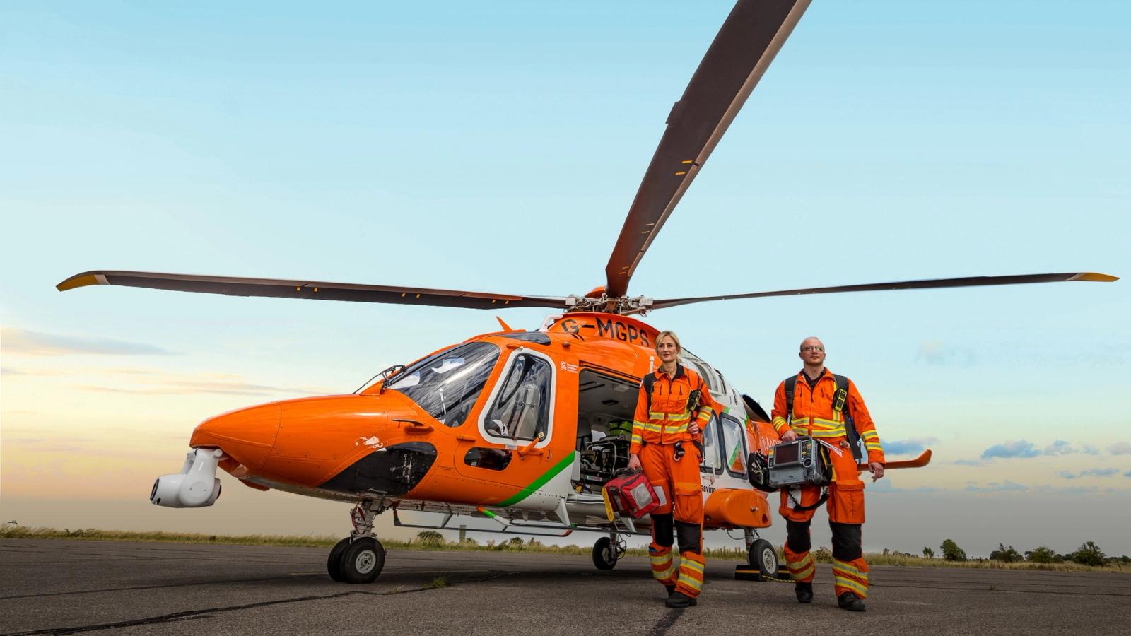 Bright orange Magpas helicopter sat on launchpad with two paramedics walking away from the helicopter in highly visible orange jumpsuits carrying emergency health care equipment.