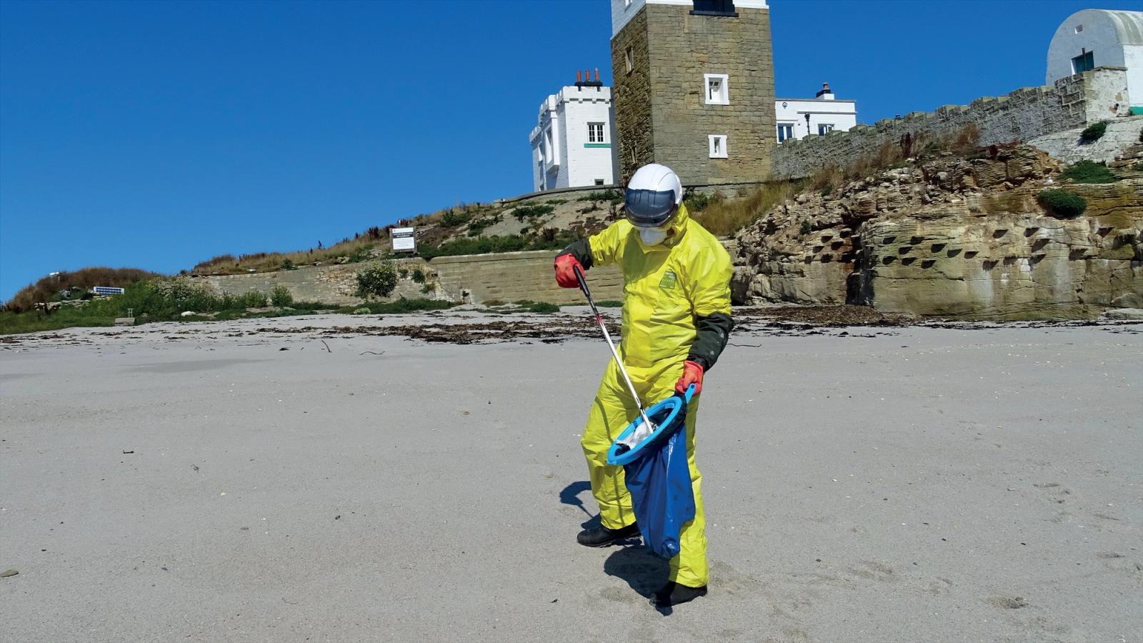 A man in a yellow hazmat suit wearing a respirator and safety equipment, cleaning items from a beach in front of a building with blue skies behind.