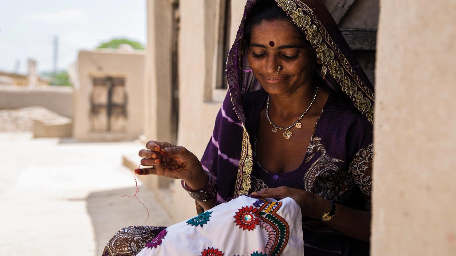 A smiling Indian woman wearing beautiful embroidered purple and gold sari sat on a step outside a sandstone house looking down sewing a piece of embroidered cloth.