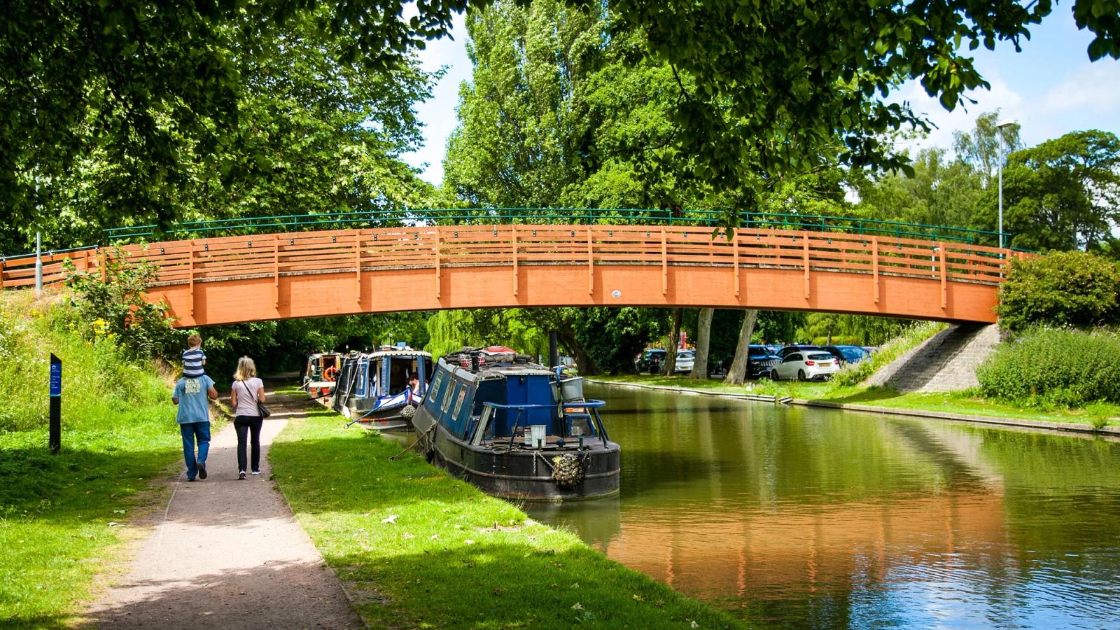 A sunny day with a family walking towards a bridge spanning a canal on a canal path past barges that are moored to the side.