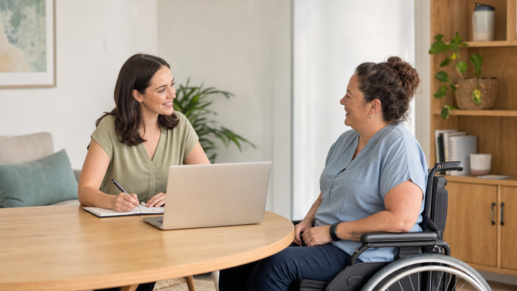 An NDIS participant using a wheelchair smiles while discussing her support needs with a coordinator at a table in a bright, welcoming office.