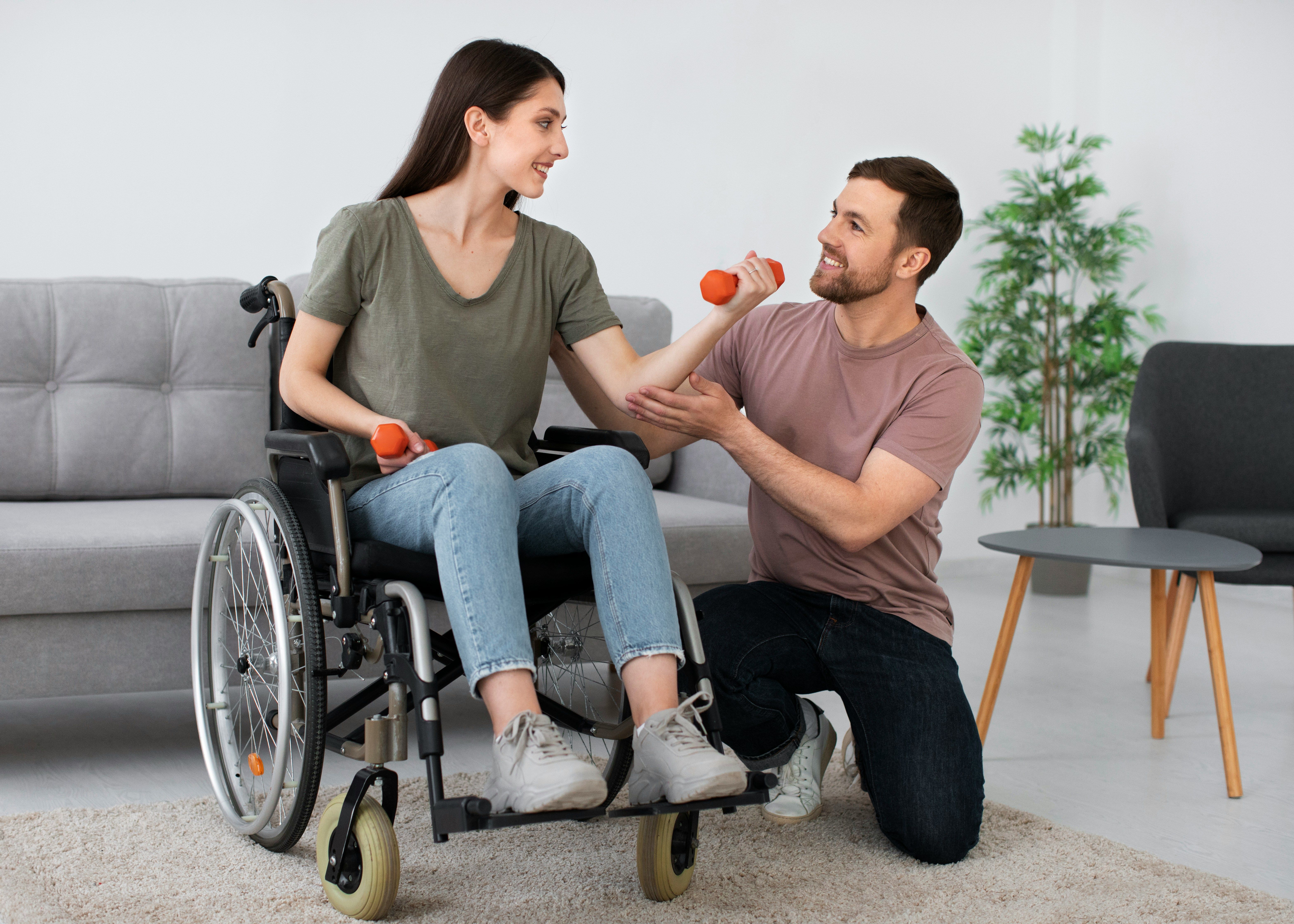 A wheelchair user performs arm exercises with dumbbells while a support worker guides her movement at home.