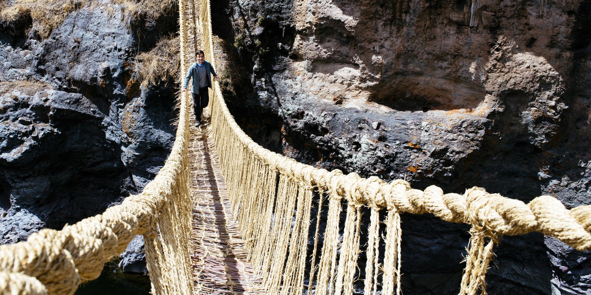 A man in a grey jacket crosses a narrow woven-grass rope bridge over a rocky gorge, representing the risk of optimising for short-term gain.
