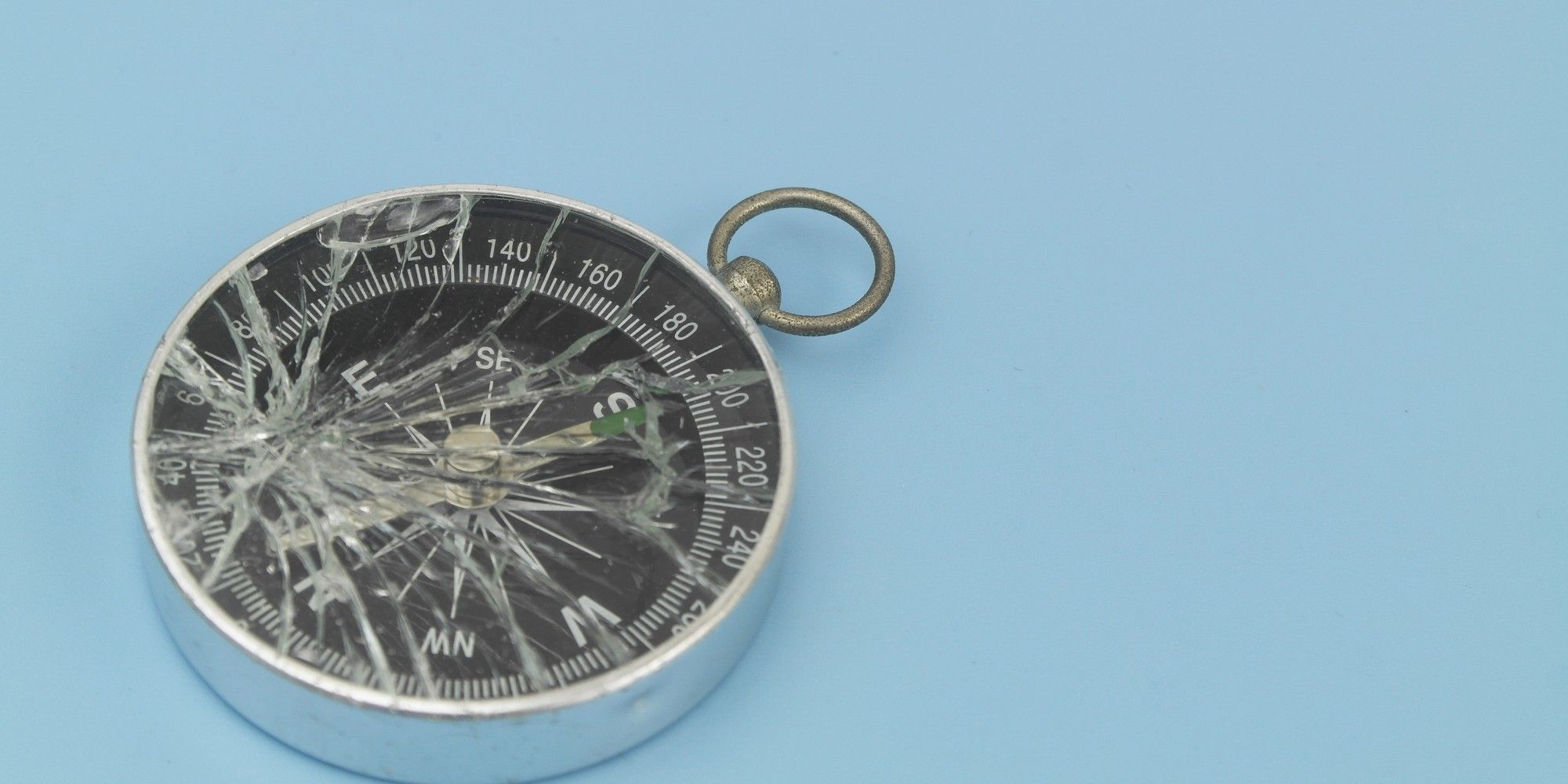 A silver pocket compass with a brass ring, its glass face shattered into spiderweb cracks, resting on a pale blue surface, symbolising misdirected attribution.