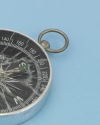 A silver pocket compass with a brass ring, its glass face shattered into spiderweb cracks, resting on a pale blue surface, symbolising misdirected attribution.