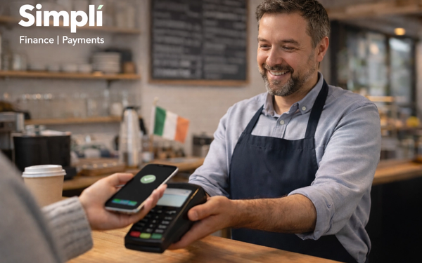 A smiling small café owner stands behind a wooden counter while holding out a card terminal as a customer taps a smartphone to make a contactless payment. The setting is a warm, modern café with shelves, a menu board, takeaway coffee cup, and a small Irish flag visible in the softly blurred background.
