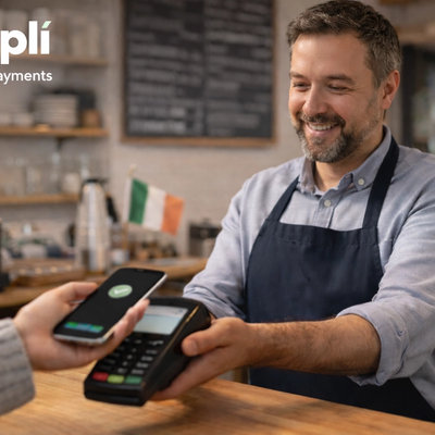 A smiling small café owner stands behind a wooden counter while holding out a card terminal as a customer taps a smartphone to make a contactless payment. The setting is a warm, modern café with shelves, a menu board, takeaway coffee cup, and a small Irish flag visible in the softly blurred background.