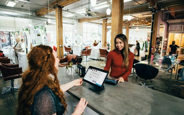 Client smiles at a modern salon reception counter while a staff member checks bookings on a laptop.