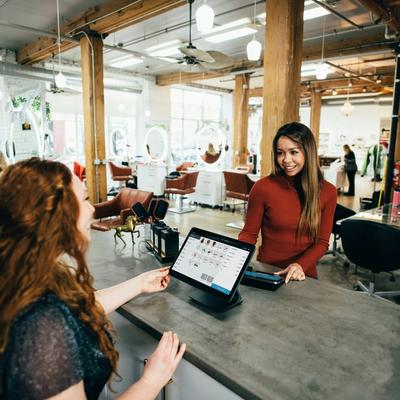 Client smiles at a modern salon reception counter while a staff member checks bookings on a laptop.