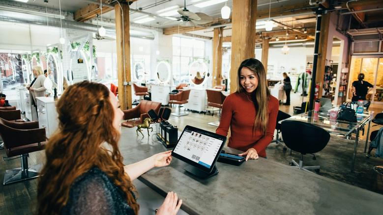 Client smiles at a modern salon reception counter while a staff member checks bookings on a laptop.