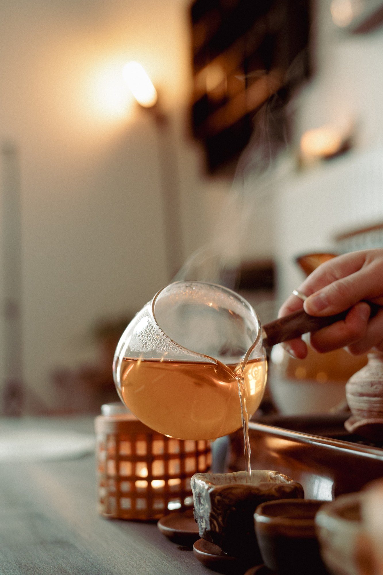 Üvegkancsóból tea csorog egy kerámia csészébe, a háttérben gyertyafény.