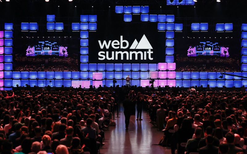 Palco principal de uma conferência de tecnologia com plateia lotada e telões iluminados, representando o Web Summit Rio 2026 no Riocentro.