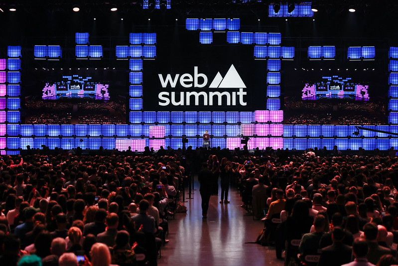 Palco principal de uma conferência de tecnologia com plateia lotada e telões iluminados, representando o Web Summit Rio 2026 no Riocentro.