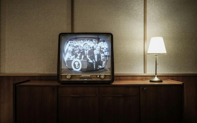 Televisor vintage dos anos 1950 ligado sobre um móvel de madeira, exibindo uma transmissão em preto e branco, com clima de época.