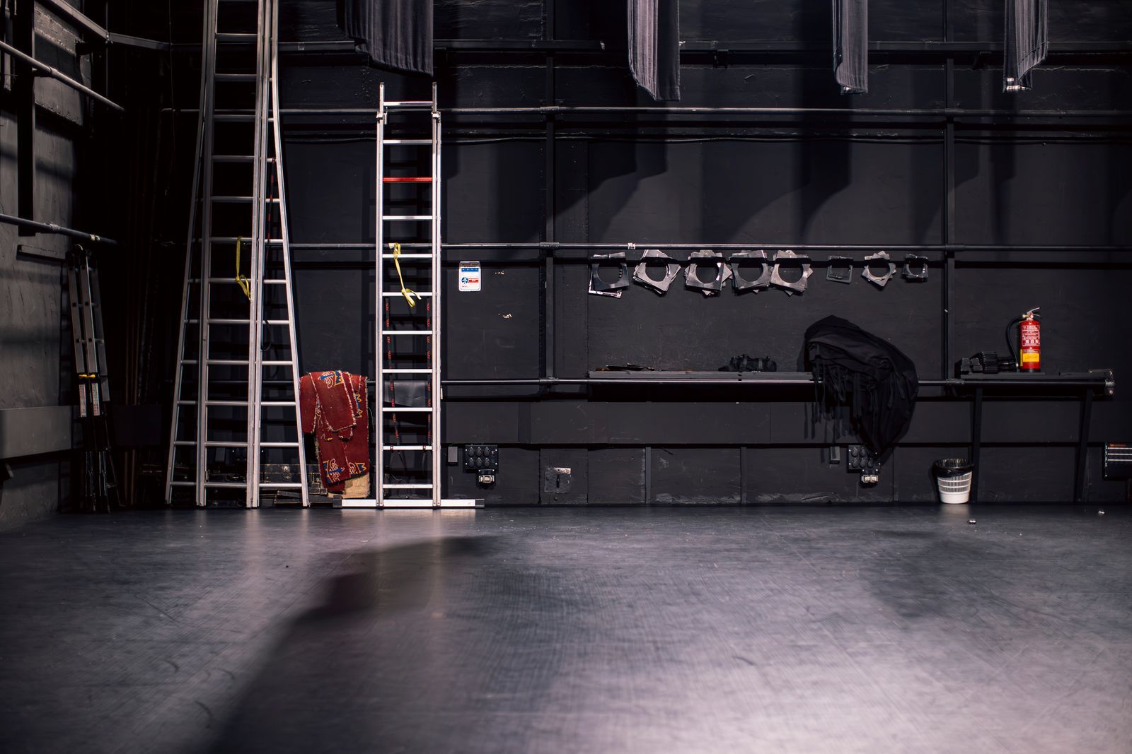 Palco de teatro vazio nos bastidores, com escadas e refletores guardados em um rack na parede, pronto para a próxima montagem de cena