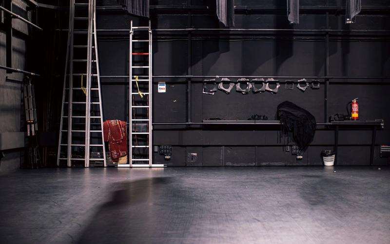 Palco de teatro vazio nos bastidores, com escadas e refletores guardados em um rack na parede, pronto para a próxima montagem de cena
