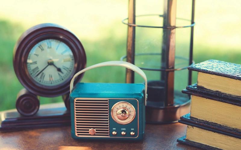 Rádio retrô azul e um relógio de mesa antigo sobre uma superfície de madeira, com livros e uma lanterna ao fundo, em cena ao ar livre iluminada por luz natural quente.