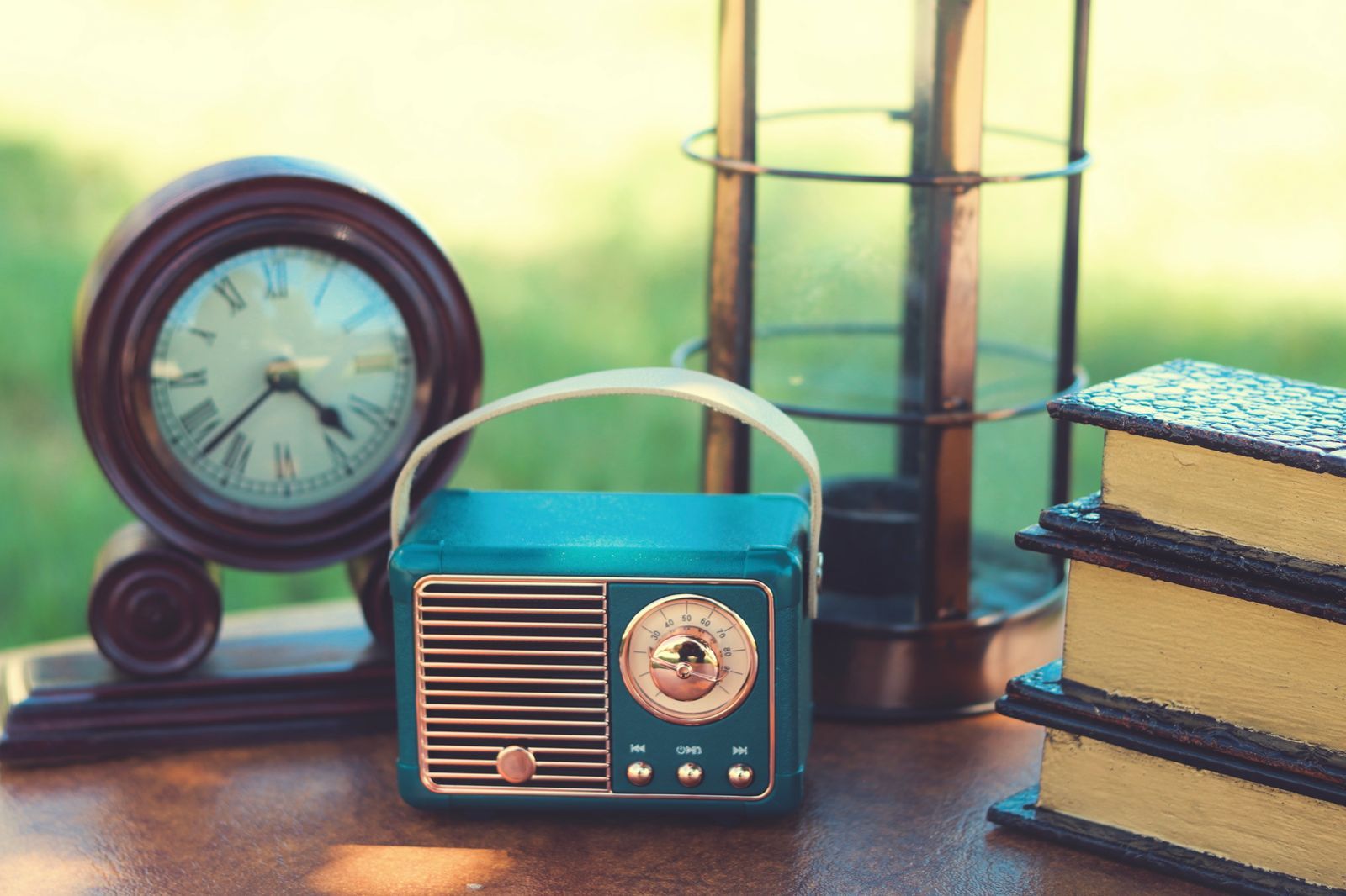 Rádio retrô azul e um relógio de mesa antigo sobre uma superfície de madeira, com livros e uma lanterna ao fundo, em cena ao ar livre iluminada por luz natural quente.