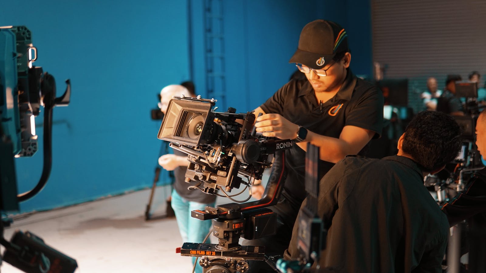 Profissional operando uma câmera de cinema em um estúdio de produção, com a equipe ao fundo — a formação prática do curso de Produção Profissionalizante da Escola de Rádio.