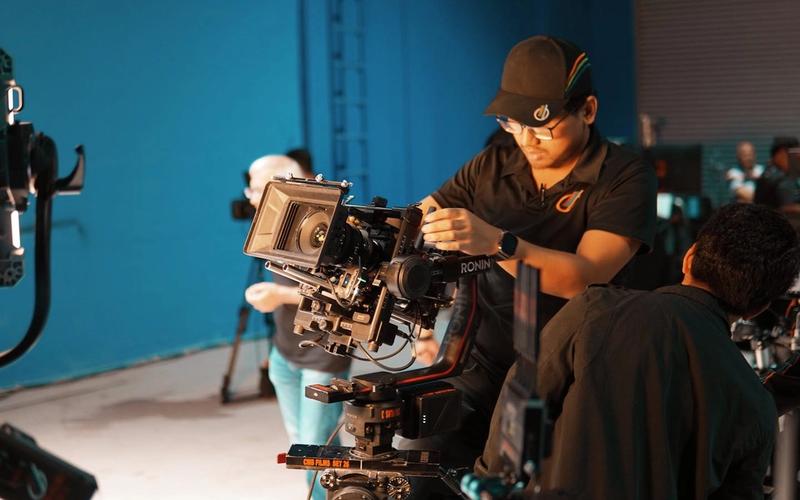 Profissional operando uma câmera de cinema em um estúdio de produção, com a equipe ao fundo — a formação prática do curso de Produção Profissionalizante da Escola de Rádio.