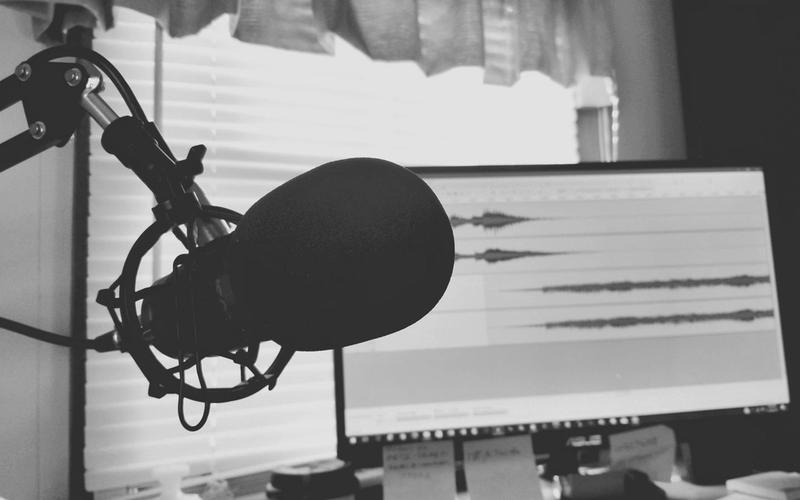 Microfone condensador em close, em preto e branco, com uma tela ao fundo exibindo formas de onda de áudio em edição, sugerindo o processo de narração e produção de um podcast investigativo.