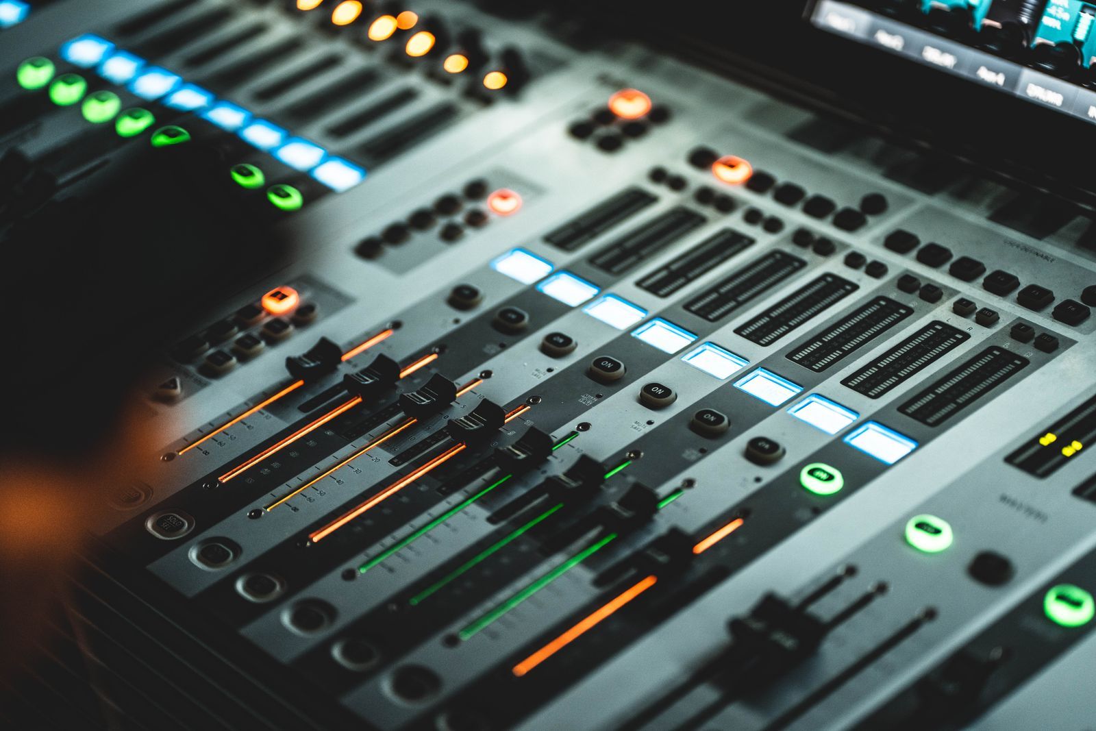 Close-up de uma mesa de mixagem digital com faders e LEDs coloridos acesos (azul, verde e laranja) em ambiente escuro, sugerindo um estúdio de rádio operando sozinho durante a madrugada.