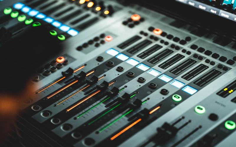 Close-up de uma mesa de mixagem digital com faders e LEDs coloridos acesos (azul, verde e laranja) em ambiente escuro, sugerindo um estúdio de rádio operando sozinho durante a madrugada.